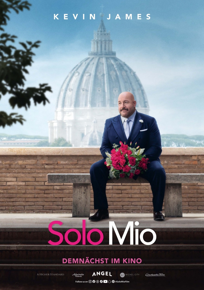 Kevin James im Hochzeitsdress mit Blumenstrauß auf einer Bank vor einer Mauer - im Hintergrund eine Kuppel - Solo Mio